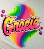 Gracie in pink-and-yellow airbrushed block script with dark outlines, pink bubbles and a rainbow swirl on white fabric.