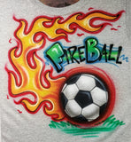 Fireball lettering above a shaded soccer ball with curling orange and yellow flames, red glow and green grass on a gray shirt.