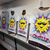 Four hand-airbrushed sunshine name shirts displayed on studio easels, showing smiling golden suns in sunglasses with colorful script names on white and gray garments.