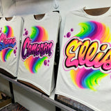Three personalized rainbow-swirl airbrush shirts displayed together on studio easels, showing different name-lettering colors for a coordinated group.
