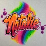 Natalie in orange-and-pink airbrushed block script with purple outlines, pink bubbles and a rainbow swirl on white fabric.