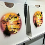 Two personalized golden sunset and palm-tree shirts displayed side by side on studio easels, showing the shoreline design with different script names.