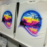 Two Pedro and Leslie sunset shirts displayed on easels, showing a matching-pair example of the personalized beach design; shirts ordered separately.