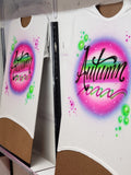 Two matching white shirts airbrushed with Autumn in black script, pink and purple speckled glow, lime green bubbles and swirls, shown together on painting boards.