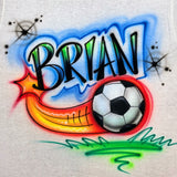 Hand-airbrushed Brian name in black lettering with blue and green highlights above a flying soccer ball, orange and yellow motion trail, starbursts and green grass accents on light fabric.