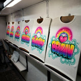 Six personalized airbrush shirts on studio easels, each with a different name beneath a rainbow and pink heart, framed by turquoise clouds.