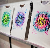 Three hand-painted hibiscus name shirts on the studio easel, showing green and turquoise, purple and blue, and pink, yellow and turquoise color variations.