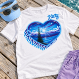 Kathy nighttime heart airbrush shirt with a small script name above the blue and purple beach artwork, styled with a sky-blue visor and lavender shorts; digital mockup.