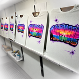 Easel lineup of personalized sunset-filled graffiti name shirts, including Natalie, with the studio background softly faded at the corners.