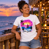 Digital mockup of a woman wearing the two-name sunset sailboat airbrush shirt, with palm silhouettes and pink skies, beside a beachside tiki-cafe railing.