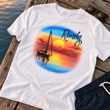 Randy golden sunset sailboat shirt on a wooden dock, with its sleeve draping over the edge beside the water; digital mockup with navy shorts and sunglasses.