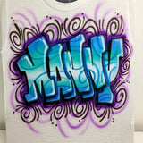 Close-up of the original hand-airbrushed Manny name design: blue and turquoise graffiti letters, black dimensional shadows and purple swirls on a white shirt.
