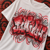 Styled mockup of a white custom-name shirt with red-and-white graffiti lettering, black shadows and swirling accents against red brick with black spray-paint swirls.