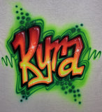 Close-up of original airbrushed Kira graffiti lettering in red, orange and yellow, with white highlights, black shadows, green outlines and dot accents on light fabric.