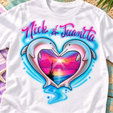 Nick and Juanita dolphin-heart shirt mockup, closer view on a beach flatlay with aqua wood and a purple towel.