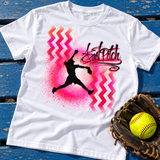 White T-shirt with red and pink chevrons, a softball pitcher silhouette and FastPitch lettering, laid on blue boards beside a glove and softball.
