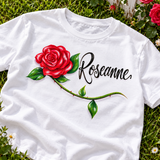 White Roseanne rose-and-script shirt angled on garden grass beside roses and a white fence; digital flatlay mockup showing the localized artwork placement.