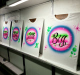Four coordinating white shirts on studio easels with Big, Little, GBig and GGBig black script over pink-and-blue oval backgrounds, green bubbles and starbursts.