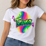 Model mockup showing the Trina rainbow swirl and green block-script name design on a white T-shirt with blue jeans.