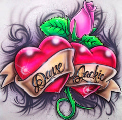 Two shaded red hearts share a flowing ribbon, framed by a pink rose, green leaves and smoky black scrollwork. Your two names replace the sample lettering across the ribbon.